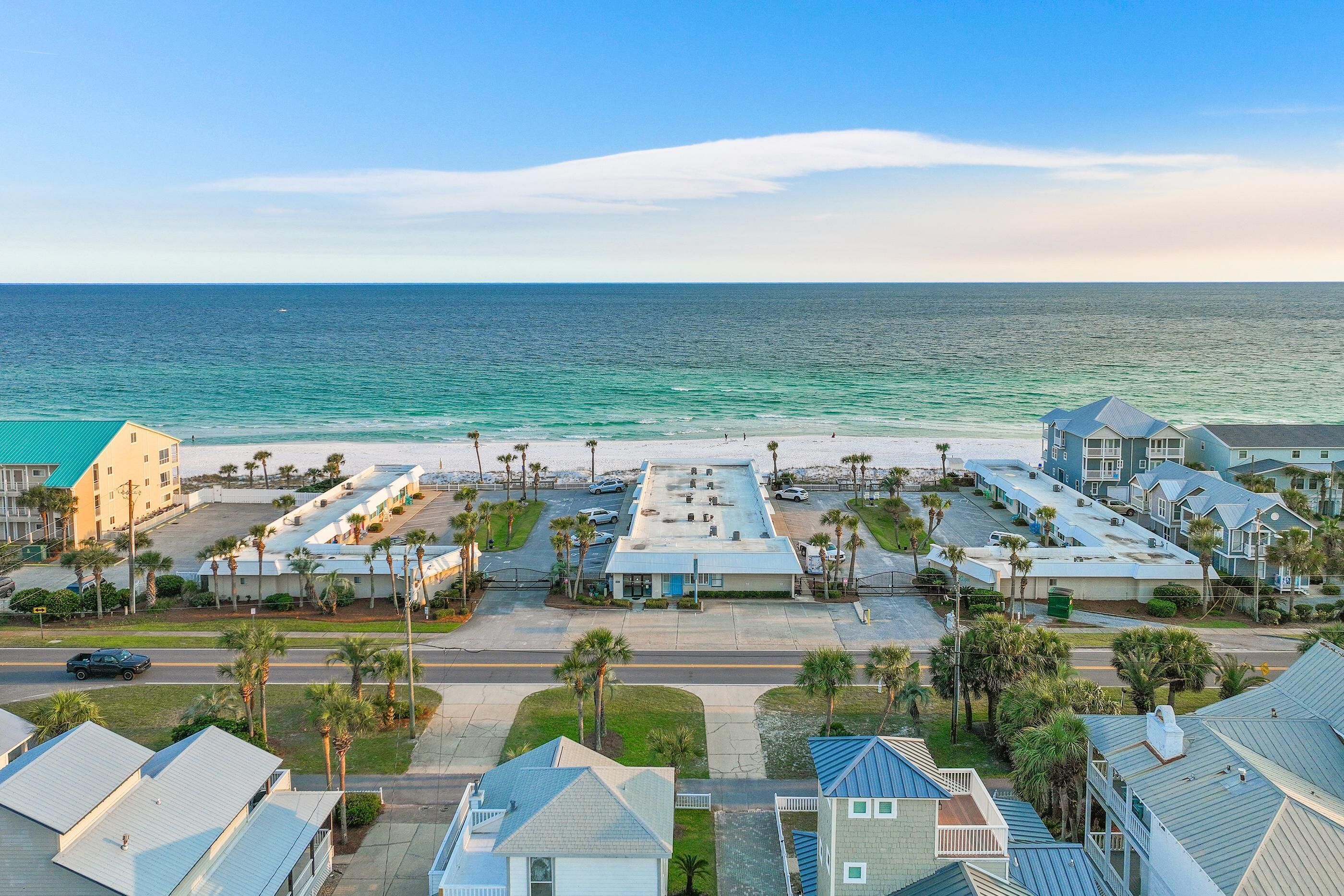 A coastal hotel with a view of the ocean, featuring a parking lot, several buildings, and palm trees.