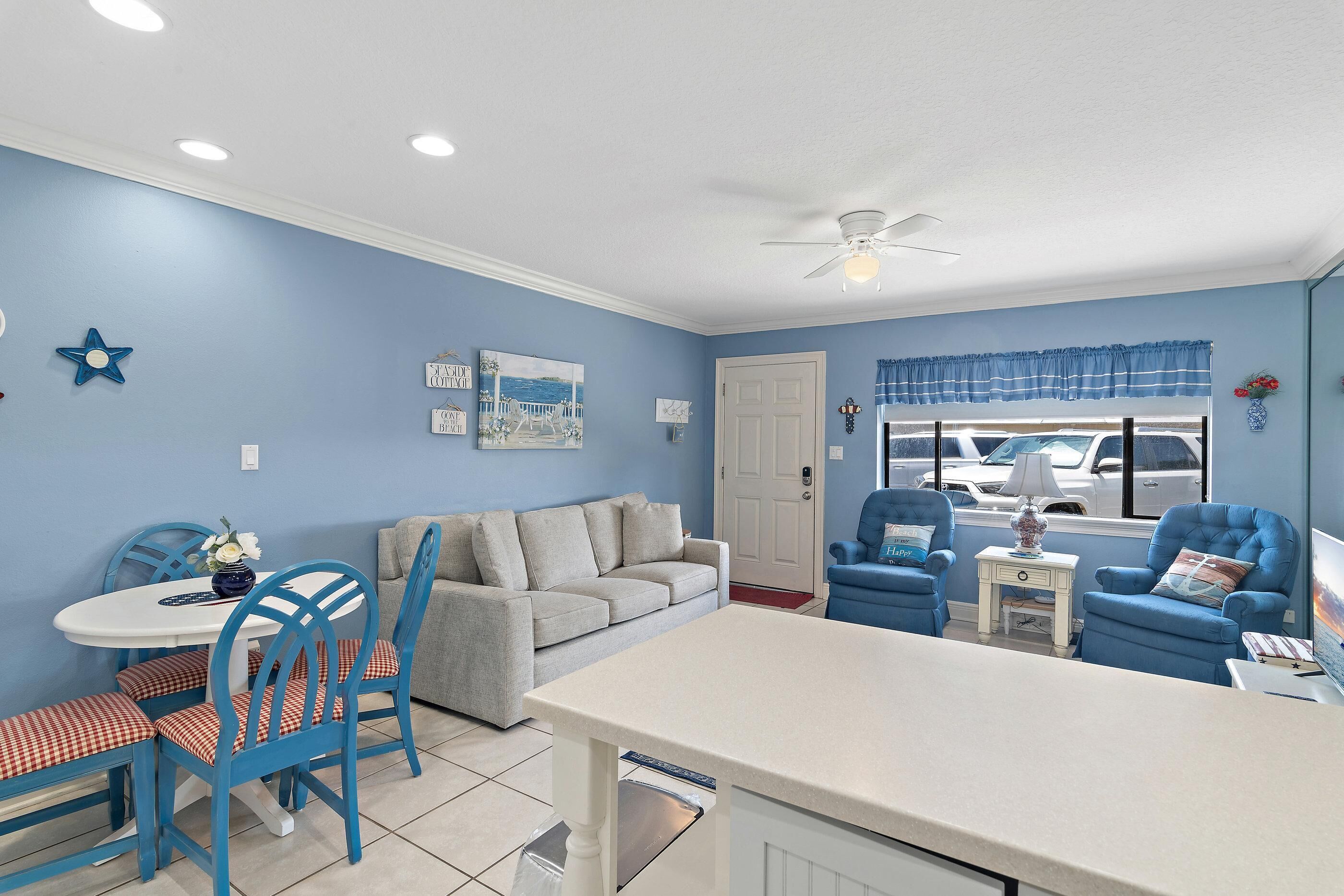 A room with a blue wall, a white ceiling fan, a grey sofa, a blue armchair, a white table with blue chairs, and a window with a view of parked cars.