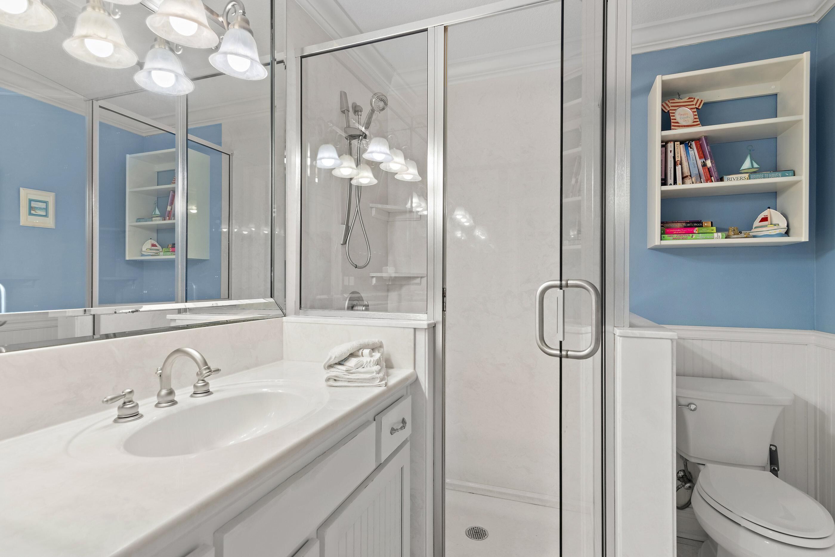 A bathroom with a white sink, mirror, and glass-enclosed shower. There is a bookshelf with books and a small decorative item.