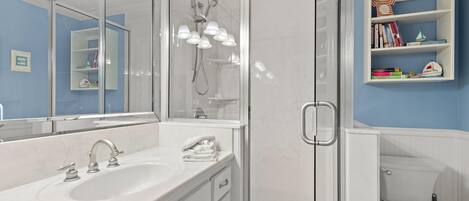 Un baño con lavamanos blanco, espejo y ducha con mampara de vidrio. Se observa una estantería con libros y un pequeño adorno.
