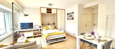 Un dormitorio moderno con cama, mesita de noche y televisor. Cuenta con un comedor con mesa y sillas.
