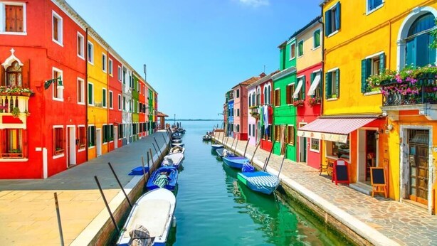 Un canal étroit bordé de bâtiments aux couleurs vives, des bateaux amarrés le long de l’eau et un ciel dégagé.