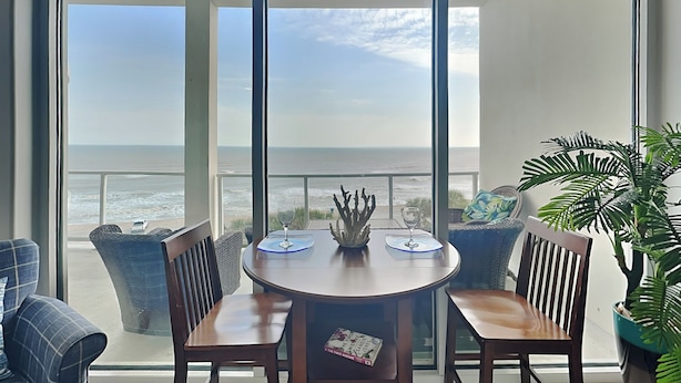 A balcony with a view of the ocean, a round wooden table with two chairs, and a potted plant. - High Seas by Ryson Vacation Rentals (Galveston)