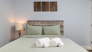 Een net opgemaakt bed met groene beddengoed en opgerolde witte handdoeken, een houten hoofdbord, een aan de muur bevestigde lamp en drie decoratieve panelen erboven.