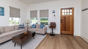 Een woonkamer met een beige bank, een houten salontafel en een bruine stoel. Er zijn drie ramen met beige jaloezieën, een houten deur en een klein schilderij aan de muur.