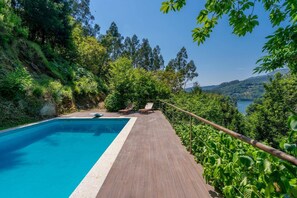 A swimming pool with a wooden deck and lounge chairs, surrounded by lush greenery and a view of a lake and hills. - Casa Azevinho - Caniçada Gêres (Vieira do Minho)