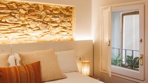 A bedroom with a stone wall headboard, a bed with white linens, and a window with a view of greenery. - Bravissimo Calderers El Cavall, in the Barri Vell (Girona)