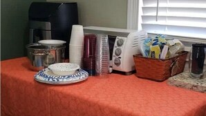 A table with a red tablecloth, a coffee maker, stacked white and pink cups, a microwave, a basket of snacks, and a plate with a blue and white design.