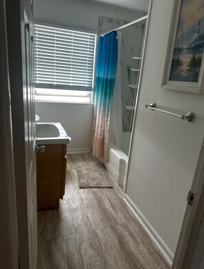 A bathroom with a shower, a window with blinds, a towel rack, and a door.