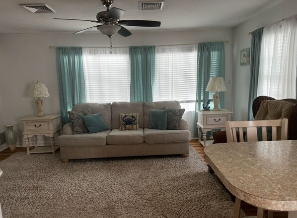 A living room with a beige sofa, two side tables, a dining table, and a ceiling fan.