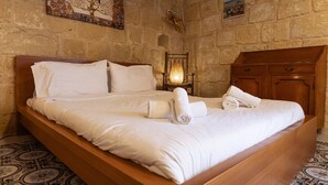 A large bed with white linens and towels, a wooden headboard, and a stone wall with a mosaic artwork. - Historic Hideaway; Stunningly Converted Studio (Ir-Rabat Għawdex)