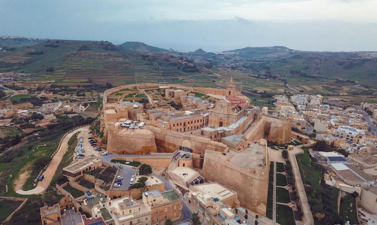 Vue aérienne d’une ville historique fortifiée, avec une imposante structure rappelant une forteresse, des rues étroites et des champs agricoles environnants.