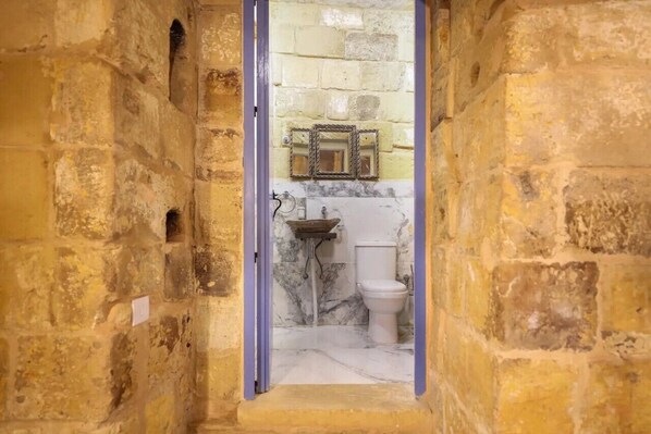 Un étroit couloir en pierre mène à une salle de bain dotée d’un lavabo en marbre et de toilettes.