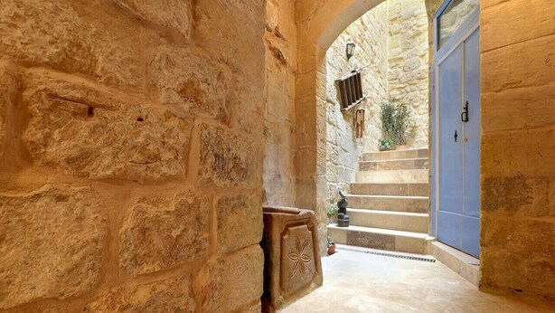 Un étroit couloir en pierre, avec une porte bleue au bout, qui mène à un escalier.