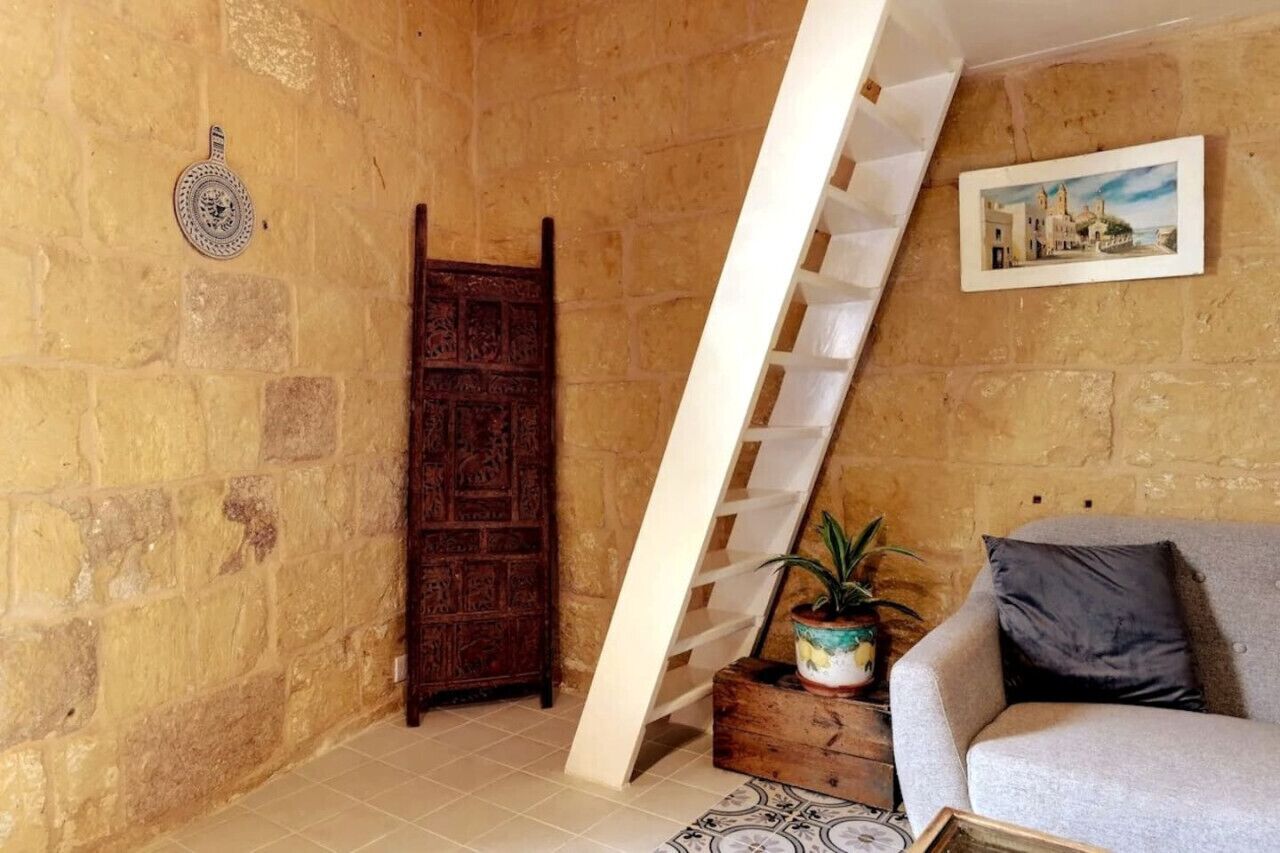 A room with a stone wall, a wooden door, a white ladder, a grey sofa, a framed picture, and a potted plant.