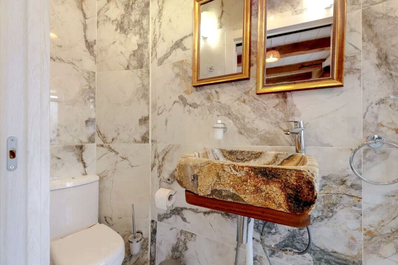 A bathroom with a marble wall, a sink with a unique stone countertop, and two mirrors with gold frames.