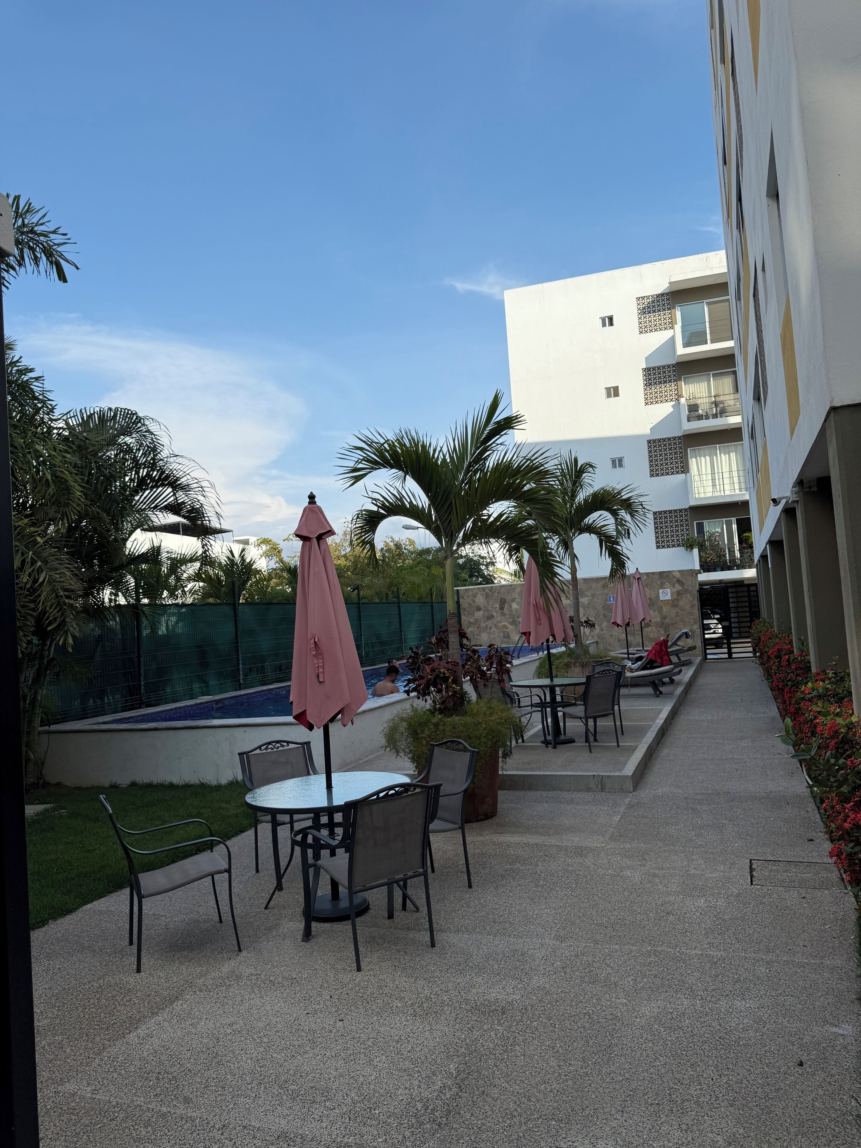 An outdoor patio area with tables and chairs, surrounded by palm trees and a swimming pool.