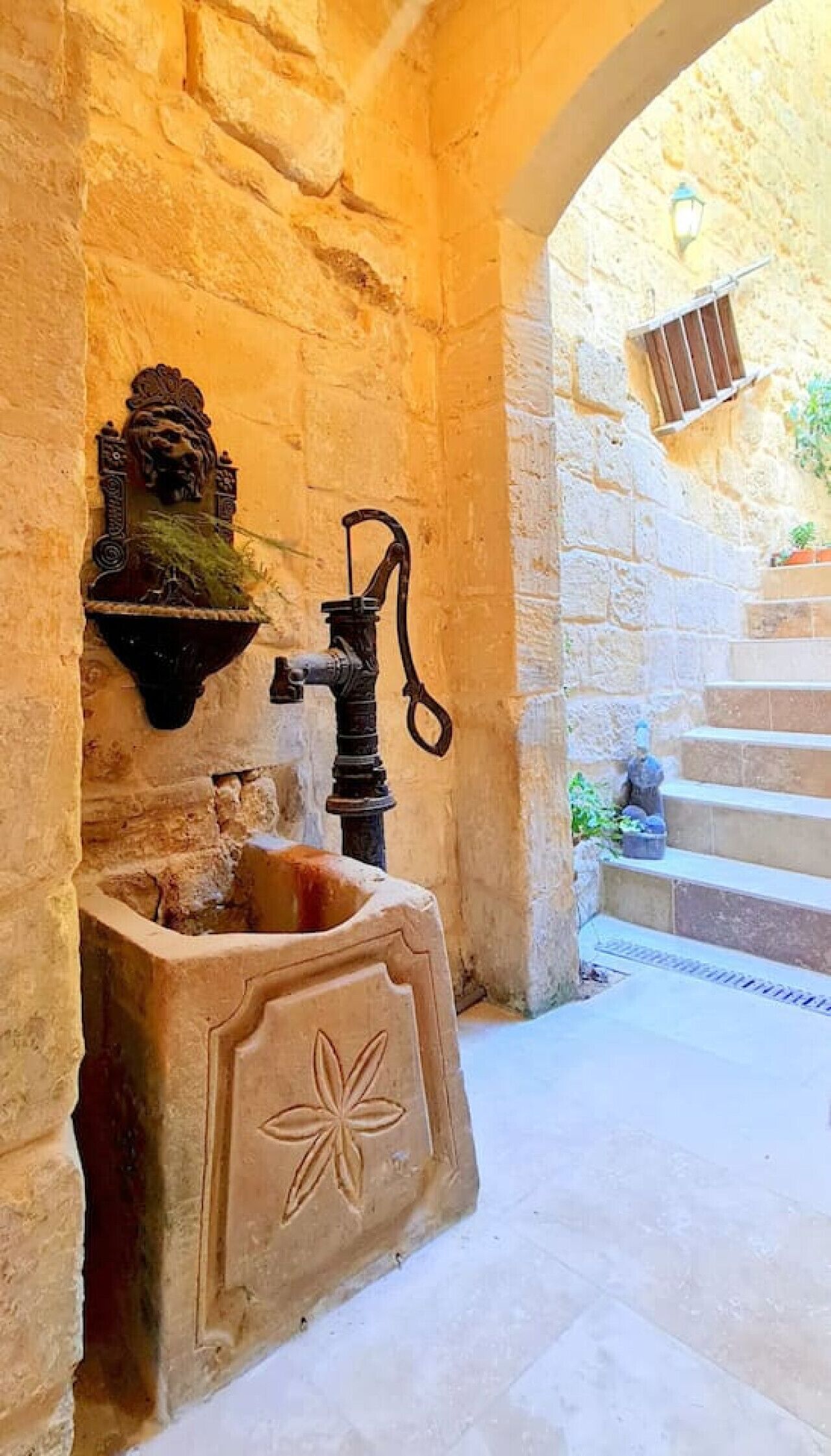 A stone well with a decorative lion head, a water spout, and a flower design on the base, set in a stone building with a staircase in the background.