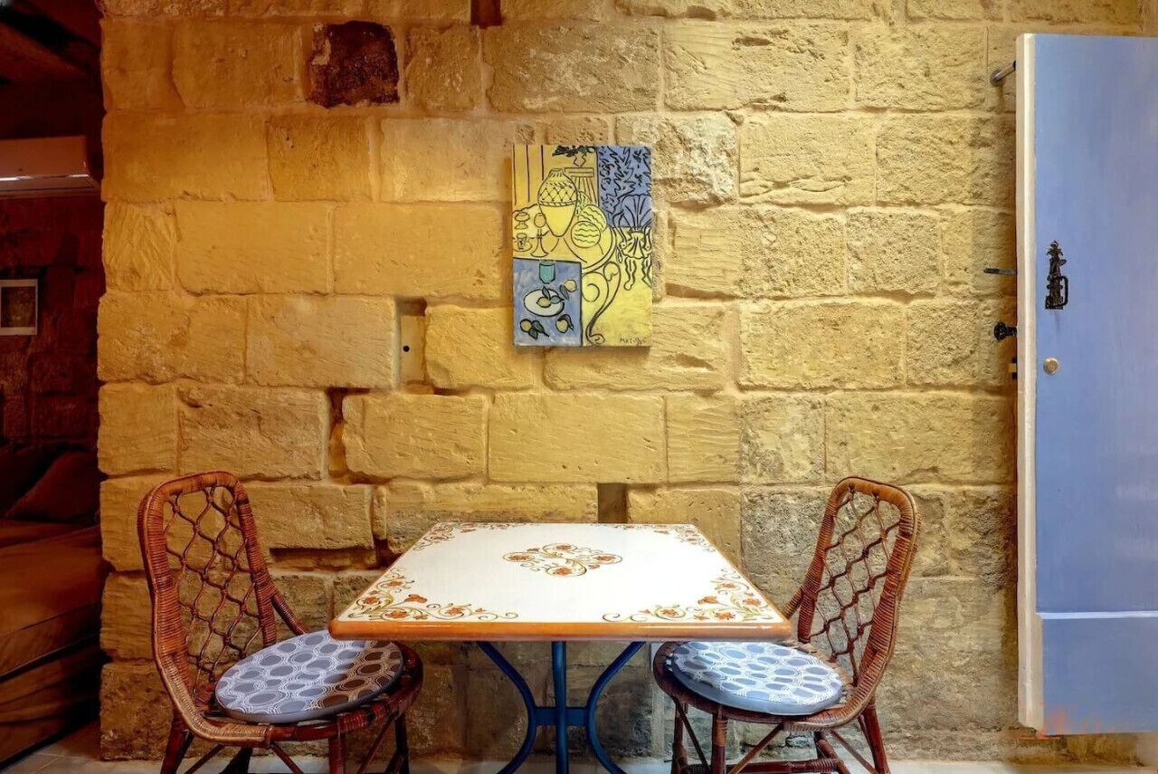 Una habitación pequeña y rústica con una pared de piedra, una mesa blanca con cubierta decorativa, dos sillas de mimbre con cojines y una puerta azul.