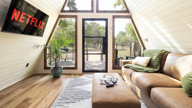 A cozy living room with a large TV screen displaying 'NETFLIX', a potted plant, a brown leather sofa, and a window overlooking a garden.