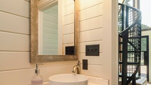 A bathroom with a white sink, a large mirror, and a black staircase visible in the background.