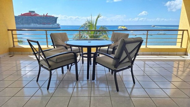 A balcony with a round table and four chairs, overlooking a body of water with a cruise ship and yachts. - Cozy oceanfront condominium-2 bed in main street, 1 block away from ferry. (Cozumel)