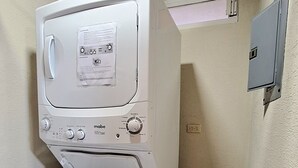 A compact, white, front-loading washing machine with a control panel, located in a small laundry room with tiled flooring and a metal rack. - Cozy oceanfront condominium-2 bed in main street, 1 block away from ferry. (Cozumel)