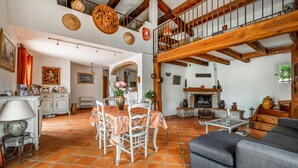 Ein geräumiges Wohnzimmer mit hohen Decken, freiliegenden Holzbalken, einem Kamin und einem Essbereich mit Tisch und Stühlen.