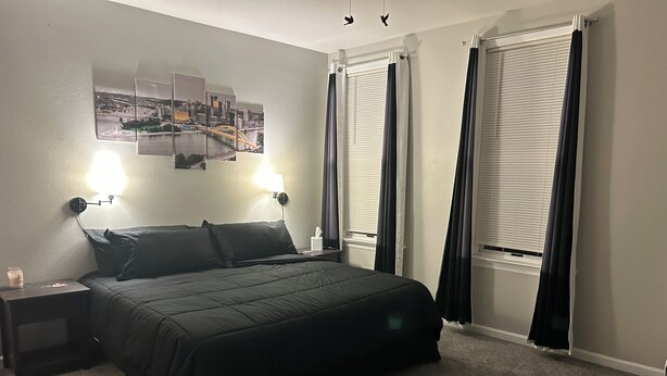 A neatly made bed with a black comforter, two bedside lamps, a nightstand with a candle, and a cityscape artwork on the wall.