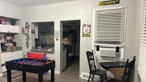 A room with a foosball table, a dining table with chairs, a kitchen area, and a window with blinds.