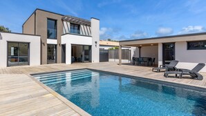 Ein modernes Haus mit Swimmingpool, einer Terrasse mit Gartenmöbeln und einer großen Glasdoor, die zu einem überdachten Außenbereich führt.
