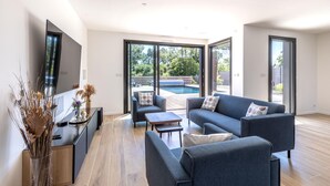 Ein modernes Wohnzimmer mit einer Couch, einem Couchtisch und einem Fernseher, große Schiebeglasfenster, die zu einem Poolbereich führen.