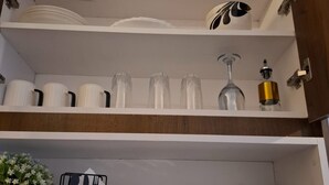 Een keukenkastje met witte planken, waarin borden, bekers, een wijnglas en een decoratief object staan. Daaronder zijn er transparante bakken met keukengerei en een kleine potplant.
