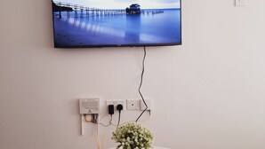 Een flatscreen-tv bevestigd aan een muur, waarop een serene zeescape te zien is. Daaronder een stekkerdoos met meerdere stopcontacten en een kleine pot met witte bloemen op een witte tafel.