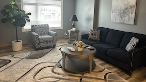Una sala de estar con un sofá azul oscuro, un sillón estampado y una mesa redonda de centro. Hay una planta en maceta, una lámpara y un cuadro en la pared.