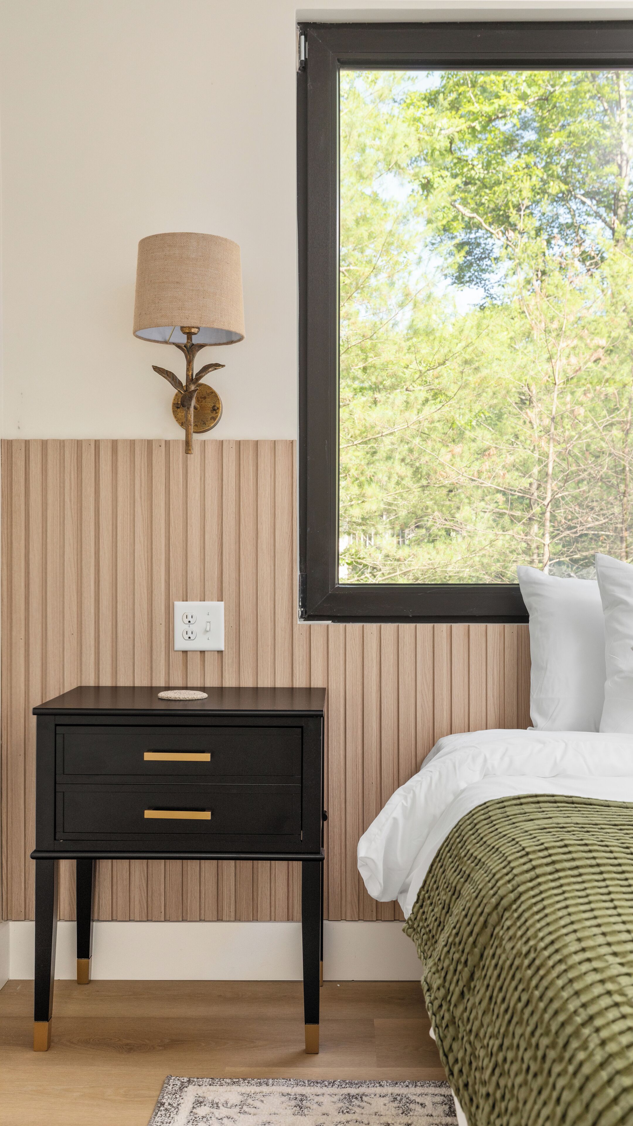 A bedroom with a bed, a nightstand with a lamp, a window with a view of trees, and a power outlet on the wall.