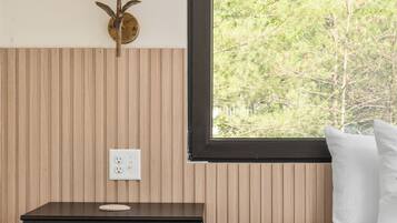 A bedroom with a bed, a nightstand with a lamp, a window with a view of trees, and a power outlet on the wall.