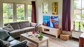 Ett vardagsrum med en soffa, ett soffbord och en tv. Det finns fönster med gardiner, ett element och ett litet bord med dekorativa föremål.
