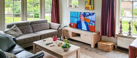 Una sala de estar con un sofá, una mesita de centro y un televisor. Hay ventanas con cortinas, un radiador y una pequeña mesa con adornos.