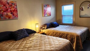 Une chambre d’hôtel avec deux lits, chacun doté d’une couette matelassée et d’oreillers à motifs. On y trouve deux tableaux accrochés au mur, une lampe sur pied et une fenêtre avec des stores.