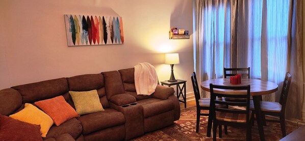 A living room with a brown sofa, colorful cushions, a small table with chairs, a lamp, and a painting on the wall. - Charming 2-bedroom apartment with equipped kitchen and AC, in Cumberland, MD. (Cumberland)