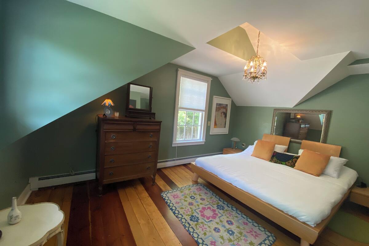 A bedroom with a sloped ceiling, a wooden dresser, a bed with white bedding, a colorful rug, and a chandelier.