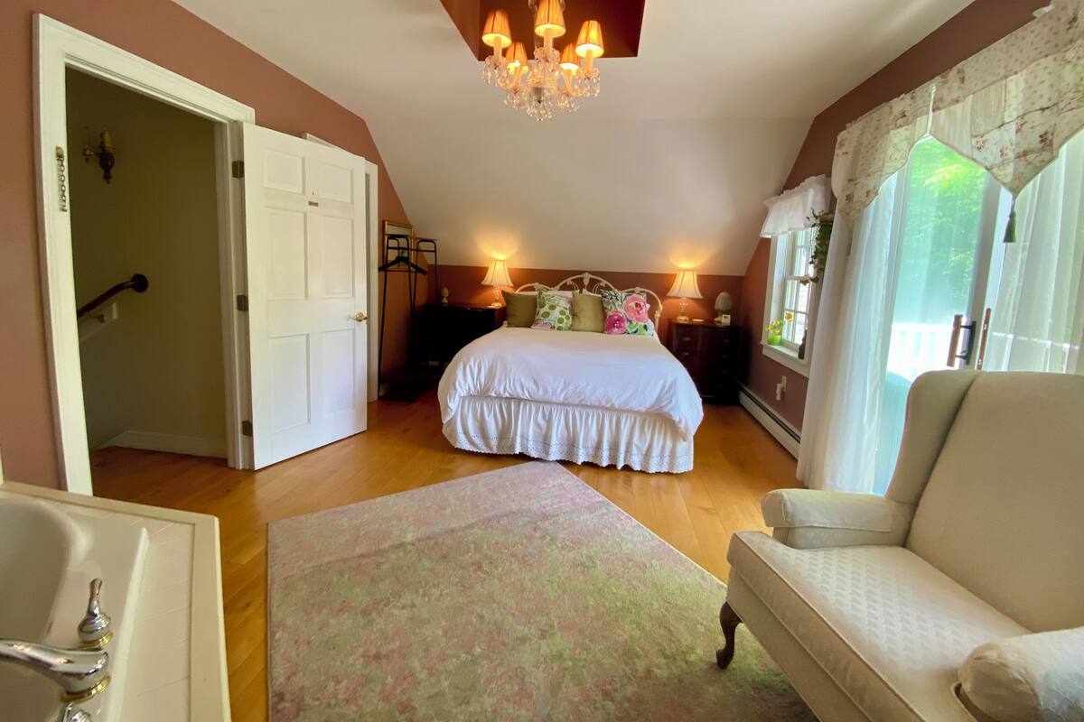 A bedroom with a bed, bedside tables, a chandelier, a window with curtains, a chair, and a door leading to another room.