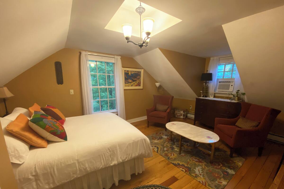 A cozy room with a bed, two armchairs, a small table, a window with a view of trees, and a ceiling light fixture.