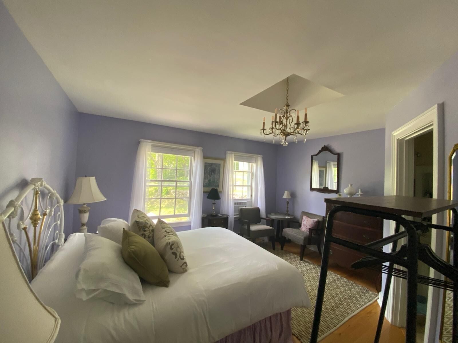 A bedroom with a bed, two chairs, a desk, a chandelier, and large windows.