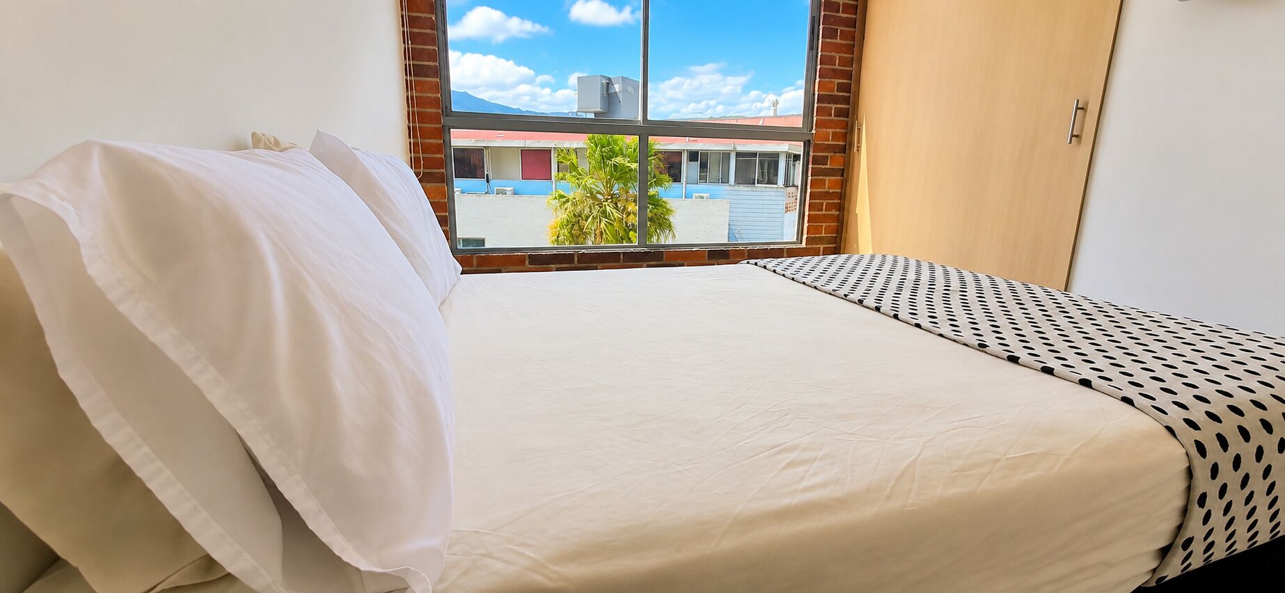A bed with white bedding and a patterned pillow, a window with a view of buildings and palm trees, and a wooden door.