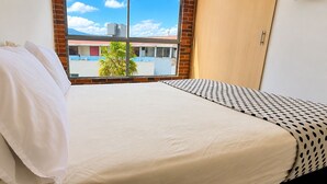 A bed with white bedding and a patterned pillow, a window with a view of buildings and palm trees, and a wooden door. - Cold AC fast Wi-Fi great location  (Medellín)