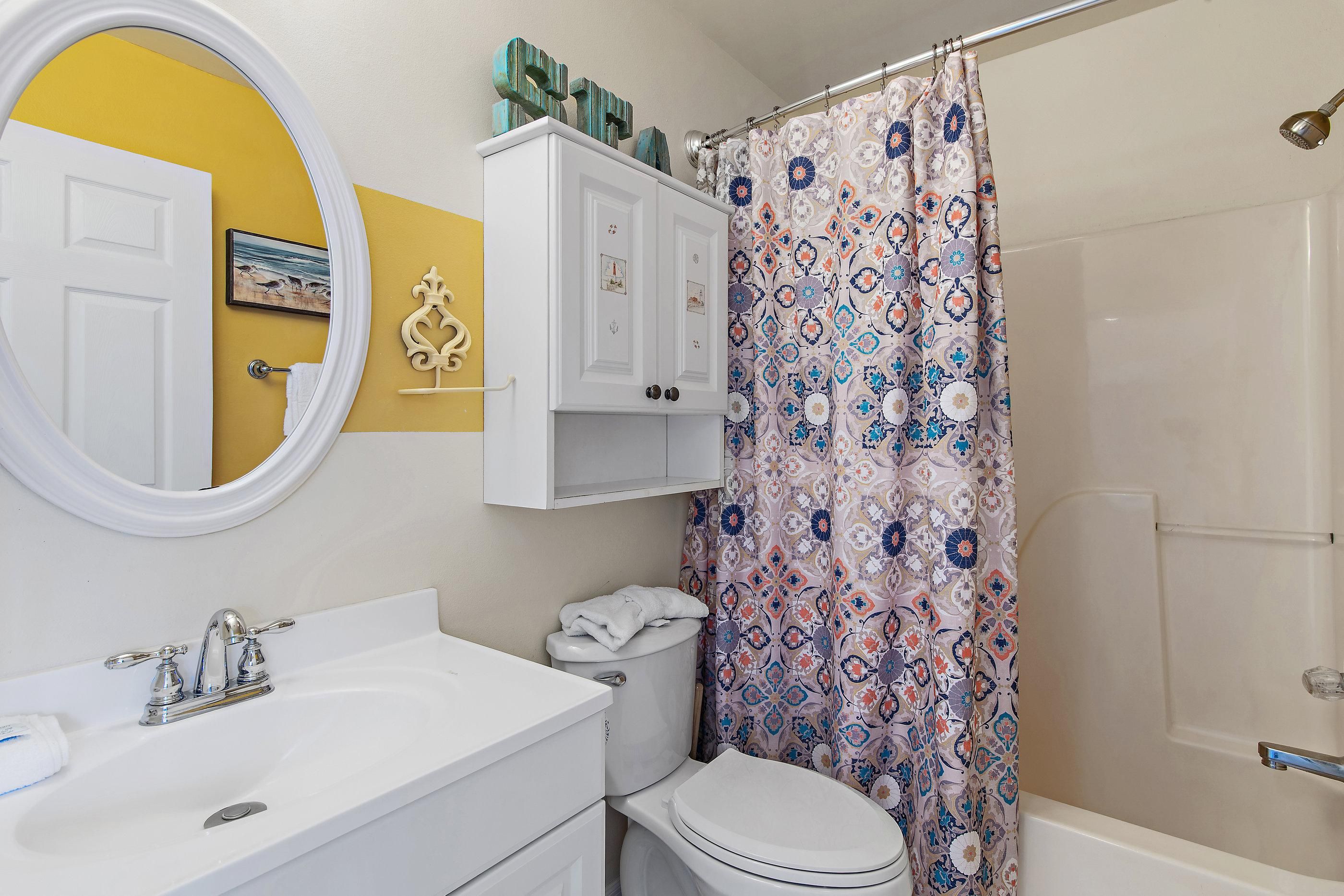 Une salle de bain avec un lavabo blanc, des toilettes, un rideau de douche à motifs floraux et un miroir devant un mur jaune.