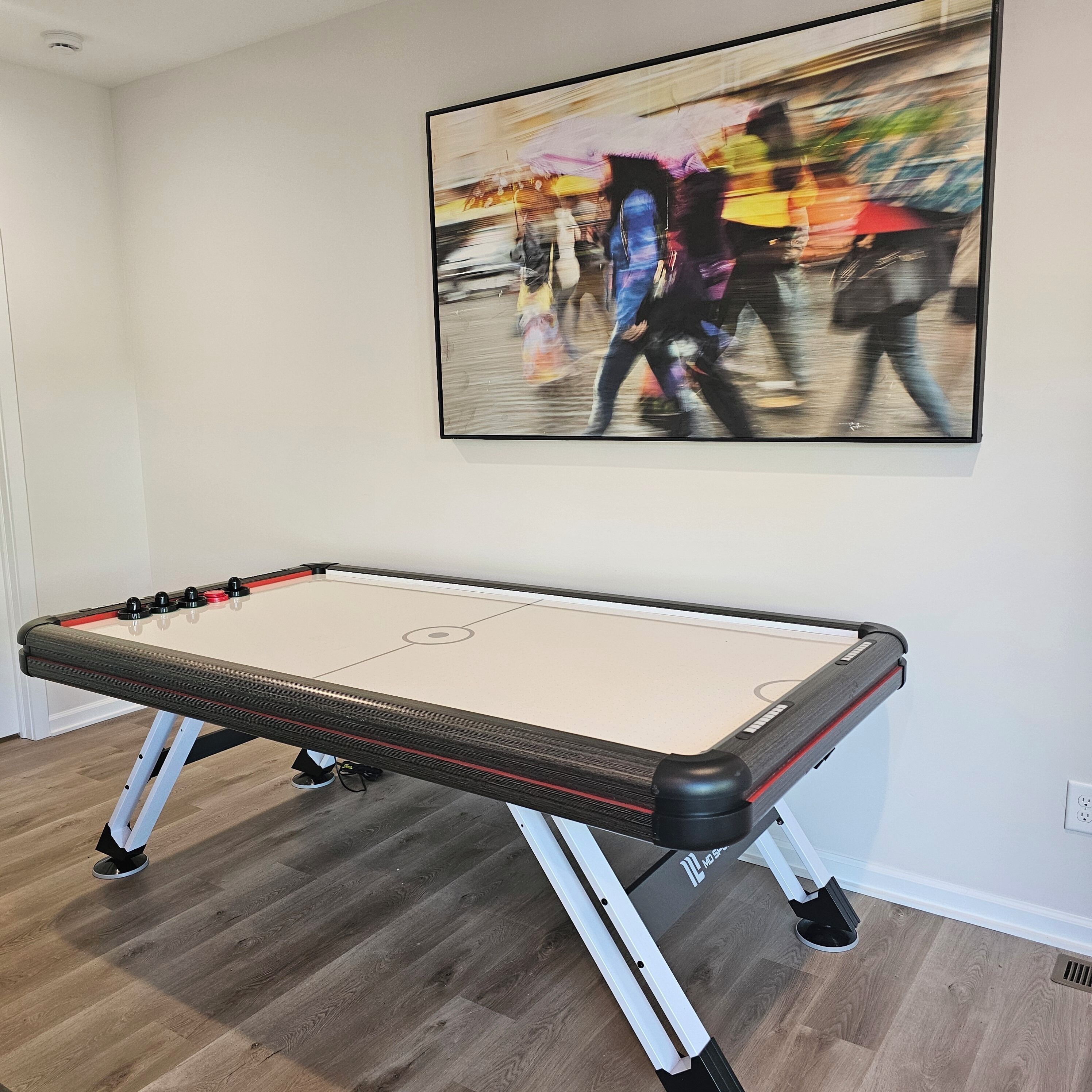 A foosball table in a room with wooden flooring and a painting of a busy street on the wall.
