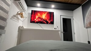 A bedroom with a bed, a mounted TV displaying a fireplace, an air conditioning unit, and a door.